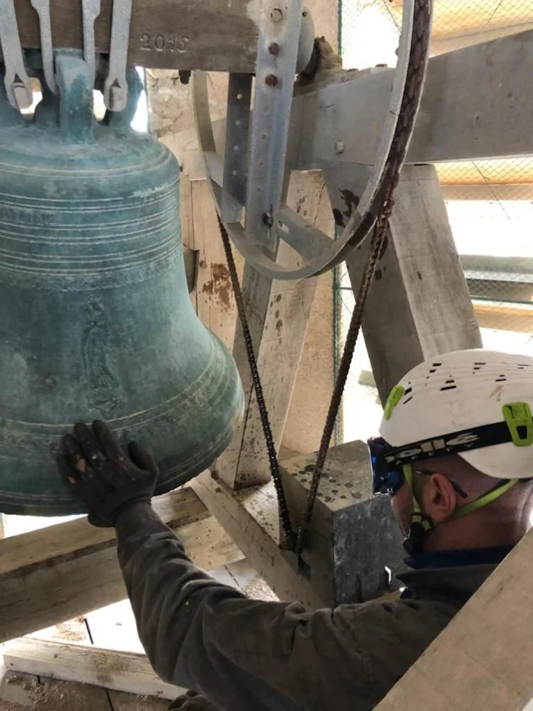 Campaniste casqué, main gantée sur une cloche patinée suspendue à son joug daté de 2016